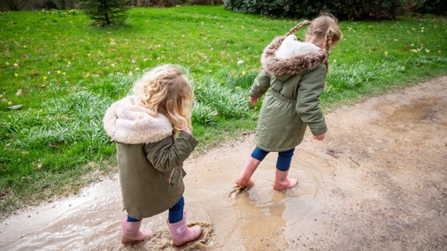Two younger girls, both wearing pink wellies, blue jeans and hooded green coats walk through big puddles on a path